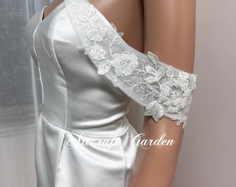 Puede incluir: Un vestido blanco de satén con hombros descubiertos con una superposición de encaje en el corpiño y un hombro. El encaje presenta delicados diseños florales.