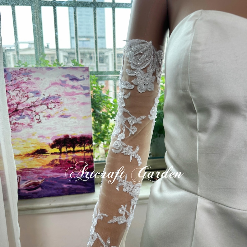 May include: A close-up of a white wedding dress sleeve with intricate floral lace detailing. The sleeve is sheer, allowing the skin to be visible. The text "Artcraft Garden" is visible on the sleeve. A painting of a sunset is in the background.