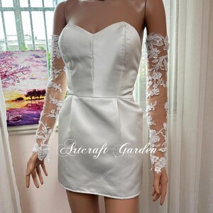 May include: A white satin strapless mini dress with a sweetheart neckline. The dress is paired with sheer, long sleeves adorned with white floral lace. The text "Artcraft Garden" is printed on the dress.