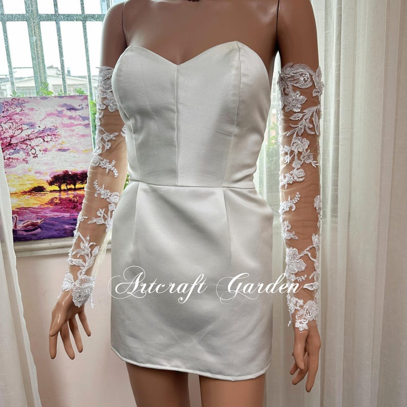 May include: A white satin strapless mini dress with a sweetheart neckline. The dress is paired with sheer, long sleeves adorned with white floral lace. The text "Artcraft Garden" is printed on the dress.