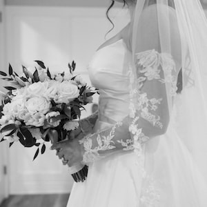 May include: A bride in a white wedding dress holds a bouquet of white roses and greenery. The dress features a strapless bodice and a flowing skirt. Delicate lace sleeves and a veil complete the bridal look. The image is in black and white.