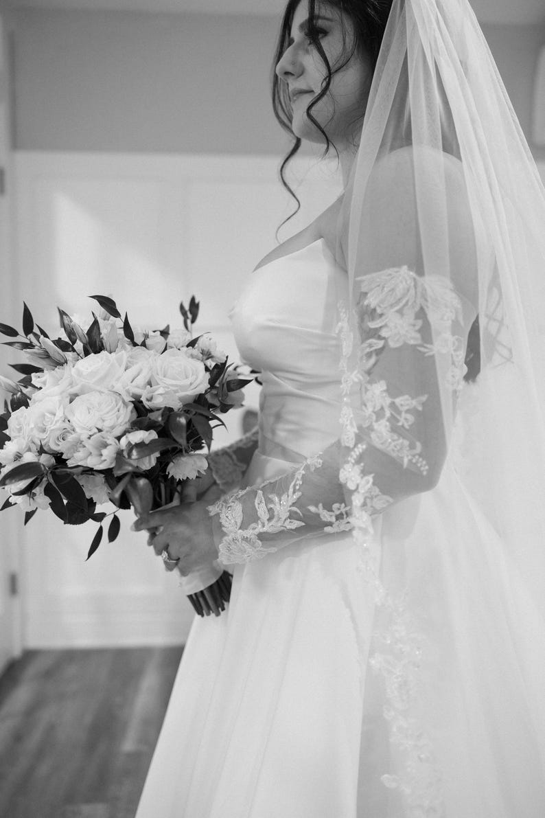 May include: A bride in a white wedding dress holds a bouquet of white roses and greenery. The dress features a strapless bodice and a flowing skirt. Delicate lace sleeves and a veil complete the bridal look. The image is in black and white.