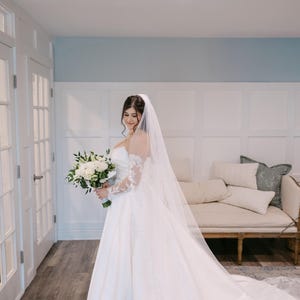 May include: A bride in a white wedding gown with a long train and lace sleeves holds a bouquet of white flowers. She wears a long veil. The background includes white doors, a light blue wall, and a cream-colored sofa.