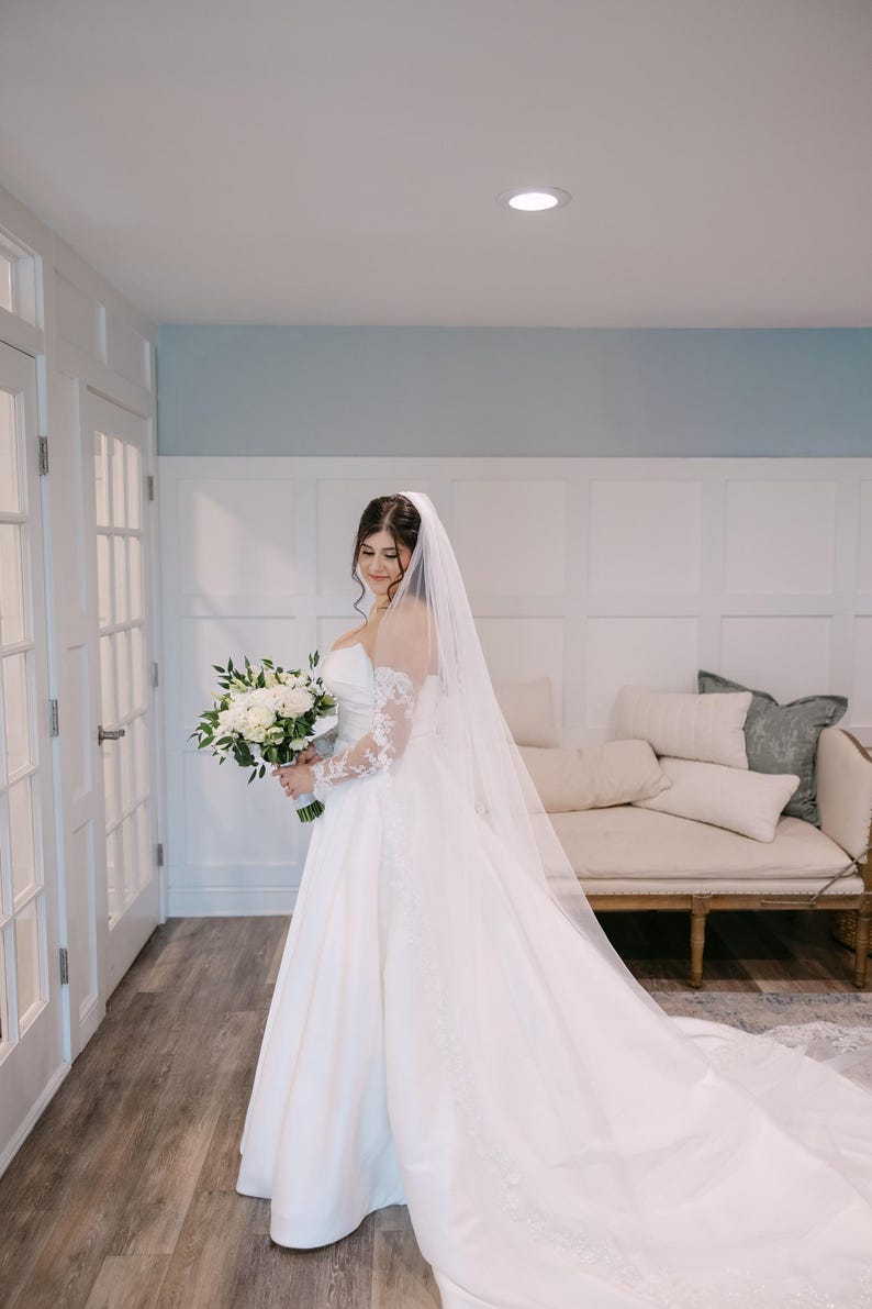 May include: A bride in a white wedding gown with a long train and lace sleeves holds a bouquet of white flowers. She wears a long veil. The background includes white doors, a light blue wall, and a cream-colored sofa.