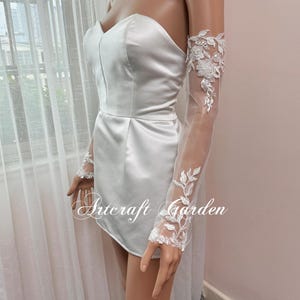 May include: White satin strapless dress with sheer lace sleeves featuring floral embroidery. The sleeves are elbow length and have a delicate, feminine look. The dress is simple and elegant, perfect for a wedding or other special occasion.