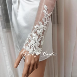 Könnte beinhalten: Weiße Braut-Handschuhe aus Spitze mit floralen Stickereien und Pailletten. Die Handschuhe sind durchsichtig und haben ein zartes Design.