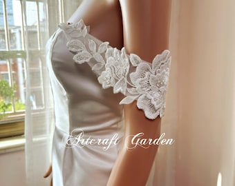 May include: White lace arm cuffs with floral embroidery. The cuffs are worn on the upper arm and feature detailed floral patterns. The text "Artcraft Garden" is visible on the dress. These cuffs are likely part of a bridal or formal outfit.