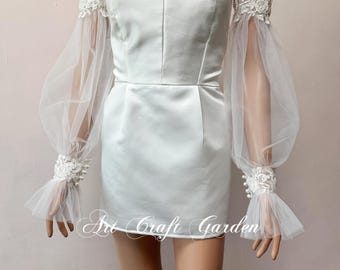May include: White off-the-shoulder dress with a sweetheart neckline and a fitted bodice. The dress has sheer tulle sleeves with lace trim and pearl embellishments. The sleeves are gathered at the wrist.