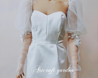 May include: White satin dress with a sweetheart neckline and puff sleeves. The sleeves are made of sheer tulle with floral embroidery. The dress has a fitted bodice and a flowing skirt. The text "Artcraft Garden" is printed on the skirt.