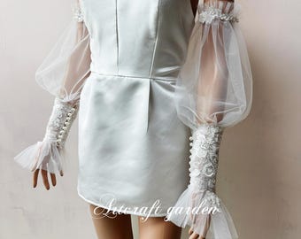 May include: White off-the-shoulder dress with a fitted bodice and a full skirt. The dress is paired with sheer white tulle sleeves with lace detailing and pearl buttons.