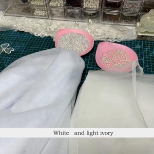 May include: White and light ivory tulle fabric, with two pink, oval-shaped containers filled with small, silver beads. The image also shows a white lace trim and a grid-patterned cutting mat. The text at the bottom reads "White and light ivory."