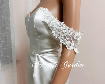 Puede incluir: Un vestido de novia de satén blanco con mangas de encaje floral. El vestido tiene un escote corazón y un corpiño ajustado. Las mangas de encaje están adornadas con flores blancas. El texto "Artcraft Garden" es visible.