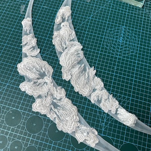 Puede incluir: Dos apliques de encaje floral blanco. Cada aplique presenta intrincados diseños florales en relieve con delicados acentos de lentejuelas. El encaje está adherido a un respaldo transparente y curvado, creando una forma elegante y fluida. Ideal para ropa de novia o adornos.