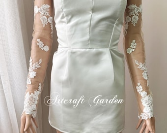 Puede incluir: Un minivestido blanco de satén con escote corazón y corpiño entallado. Mangas largas transparentes con aplicaciones de encaje floral blanco adjuntas. El texto "Artcraft Garden" está impreso en el vestido.