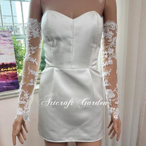 May include: A white strapless mini dress with matching sheer lace sleeves. The sleeves are adorned with floral lace appliques. The dress has a sweetheart neckline and a fitted bodice. The text "Artcraft Garden" is printed on the dress.