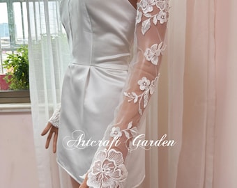 May include: White lace bridal gloves with floral embroidery. The gloves are sheer and have a delicate design. The gloves are shown on a model wearing a white strapless dress.