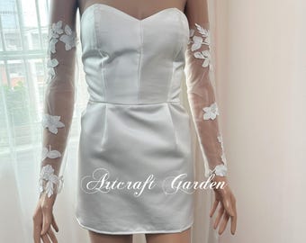 Puede incluir: Un minivestido de satén blanco con escote corazón y una superposición transparente. El vestido presenta mangas largas de tejido transparente adornadas con bordados florales blancos. El texto "Artcraft Garden" es visible.