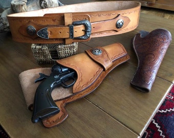May include: A light brown leather gun belt with a silver buckle and decorative star-shaped accents. A matching holster holds a black Colt revolver. A second, intricately carved leather holster is also present.
