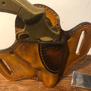 May include: A brown leather holster with a stitched design and a gun inside. The holster has a belt loop and is made of high-quality leather.