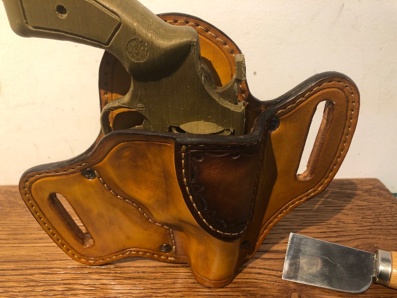 May include: A brown leather holster with a stitched design and a gun inside. The holster has a belt loop and is made of high-quality leather.