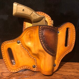 May include: A brown leather holster with a stitched design and a metal snap. A gold-colored gun is in the holster.