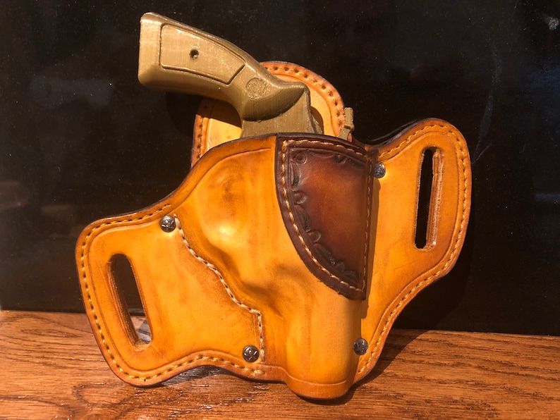 May include: A brown leather holster with a stitched design and a metal snap. A gold-colored gun is in the holster.