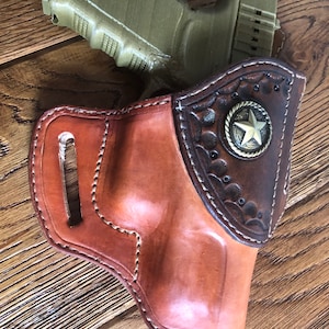 Puede incluir: Una funda de cuero marrón con un acento de metal en forma de estrella. La funda está diseñada para sostener una pistola, que es parcialmente visible en la imagen. La funda está sobre una superficie de madera.