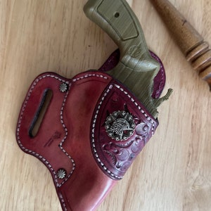 May include: A brown leather holster with a decorative eagle design and a silver star. A green plastic gun is inside the holster.