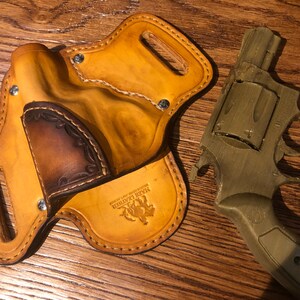 May include: A tan leather holster with a brown leather flap and a stitched design. The holster has a metal snap and the text "Jake Leather" is embossed on the bottom. A green plastic toy gun is lying next to the holster.