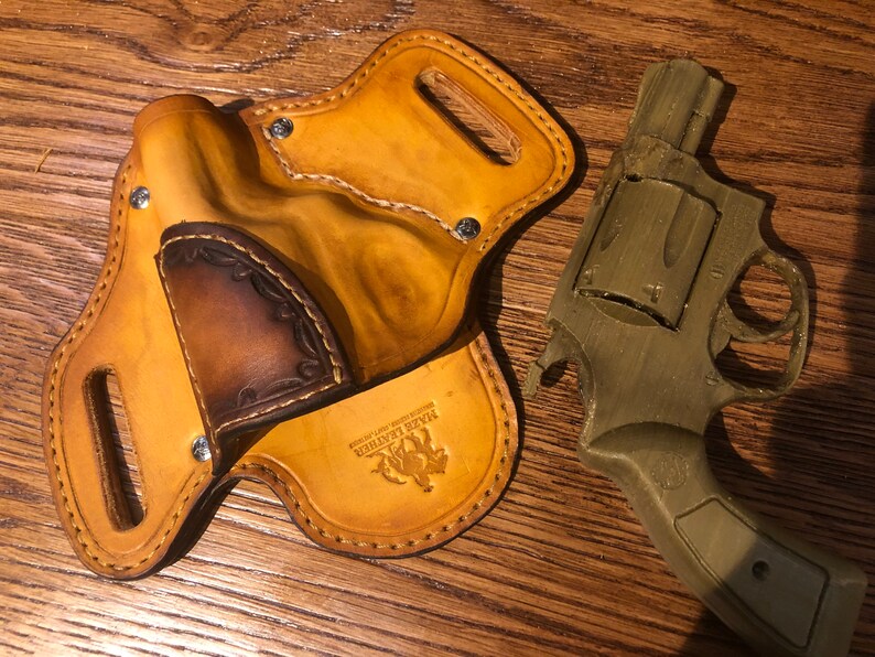 May include: A tan leather holster with a brown leather flap and a stitched design. The holster has a metal snap and the text "Jake Leather" is embossed on the bottom. A green plastic toy gun is lying next to the holster.