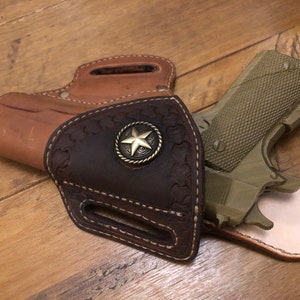 Puede incluir: Una funda de cuero marrón con un acento metálico en forma de estrella. Una pistola verde está parcialmente insertada en la funda.