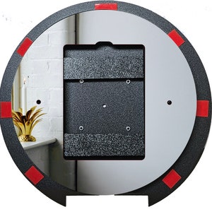 May include: A round, dark gray mirror with a textured frame and a central rectangular section. The frame has red square accents. The mirror reflects a white wall and a gold pineapple-shaped object.