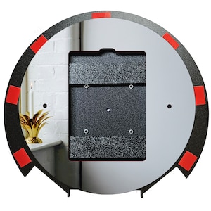May include: A round, black metal frame with a light gray interior and a rectangular, textured, dark gray insert. Red adhesive squares are placed around the frame's edge. The frame is likely for a mirror or artwork.