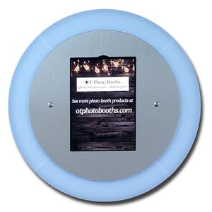 May include: A circular light blue ring light with a central gray panel featuring a black rectangular display. The display shows string lights, the text OT Photo Booths, and the website otphotobooths.com. Two small silver screws secure the panel.