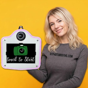 May include: A white photo booth with a pink neon border, a camera lens, and a screen displaying a green camera graphic and the words "touch to Start". The background is yellow.
