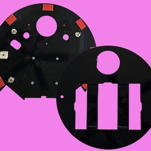 May include: Two black, semi-circular components with various holes, cutouts, and mounting hardware. Red adhesive squares are placed around the edges. One component has a large circular cutout. The background is a solid pink color.