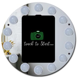 May include: A white, circular selfie light with a black rectangular screen in the center. The screen displays a green camera graphic with a smiling face and the text "touch to Start...". The light has 12 surrounding bulbs.