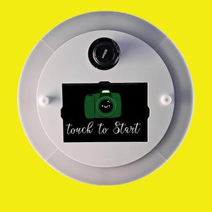 May include: A circular photo booth interface with a white and gray design. The center features a green camera icon with a smiling face on a black background, and the words "touch to Start". A Nikon lens is visible at the top.