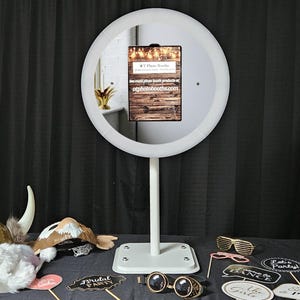 May include: A white, circular photo booth with a central screen displaying text and a wooden background. The booth is on a stand, surrounded by props like glasses and signs. Text on the screen reads "OT Photo Booths" and "otphotobooths.com".