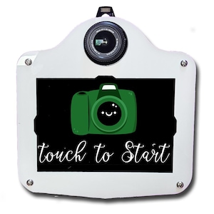 May include: White photo booth with a camera lens and a screen displaying a green camera graphic. The screen has the text "touch to Start" in white script. The photo booth has a flash and is secured with silver screws.