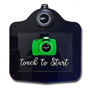 May include: Black photo booth with a camera lens at the top and a green camera graphic on the screen. The words "touch to Start" are written in white script below the graphic. The photo booth has a rounded top and a rectangular screen.