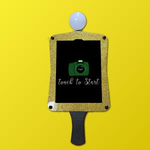 May include: A photo booth prop with a gold glitter frame and a black center. The center displays a green camera graphic with a smiling face and the words "touch to Start" in white script. A white ball is at the top.