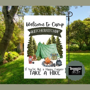 May include: A white garden flag with a camping theme. The flag features a green tent, campfire, and text that reads "Welcome to Camp QUITCHERBITCHIN' If You're Not a Happy Camper TAKE A HIKE". The flag is displayed on a black metal stand.