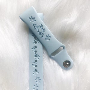 May include: Light blue watch band with puzzle piece designs and the words "Think Differently". The band is made of a soft material and has a silver clasp. The watch band is shown on a white, fluffy surface.