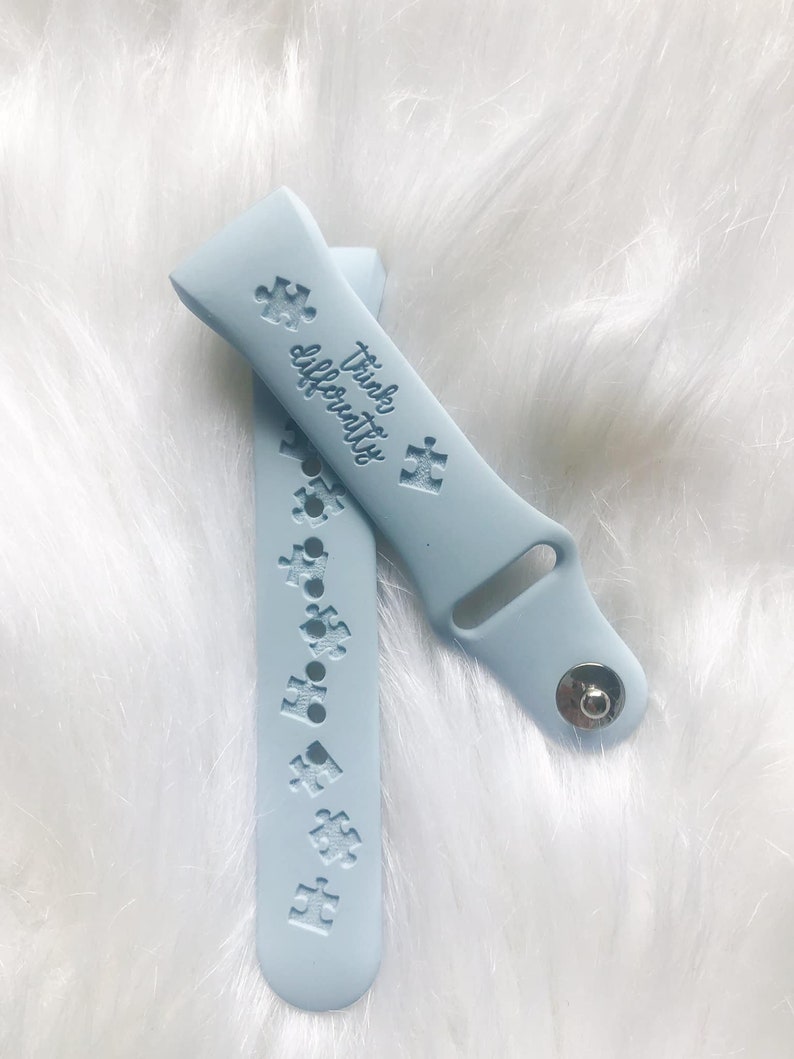May include: Light blue watch band with puzzle piece designs and the words "Think Differently". The band is made of a soft material and has a silver clasp. The watch band is shown on a white, fluffy surface.