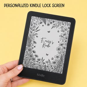 May include: A black e-reader with a personalized lock screen display featuring a black and white botanical illustration of wildflowers, ferns, and butterflies. The text "Emily's Kindle" is centered on the screen, surrounded by the floral design.