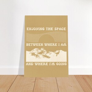 Puede incluir: Un póster de color beige con texto blanco que dice "Enjoying the space between where I am and where I'm going". El póster presenta una cordillera estilizada y un sol con rayos.