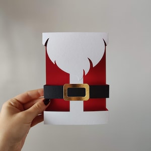 Op de afbeelding: Een witte papieren kaart met een rode en zwarte kerstman-uitsparing. De kerstman-uitsparing heeft een witte baard, een rood pak en een zwarte riem met een gouden gesp.
