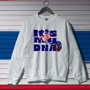 Puede incluir: Sudadera blanca con un gráfico azul y rojo que dice "It's in my DNA" con una pequeña bandera estadounidense.