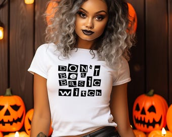 May include: A white short-sleeved t-shirt featuring the black text "DON'T BE A BASIC WITCH" printed in a ransom note style with individual letters inside black squares. The shirt is worn with black pants featuring a dark metal belt buckle.